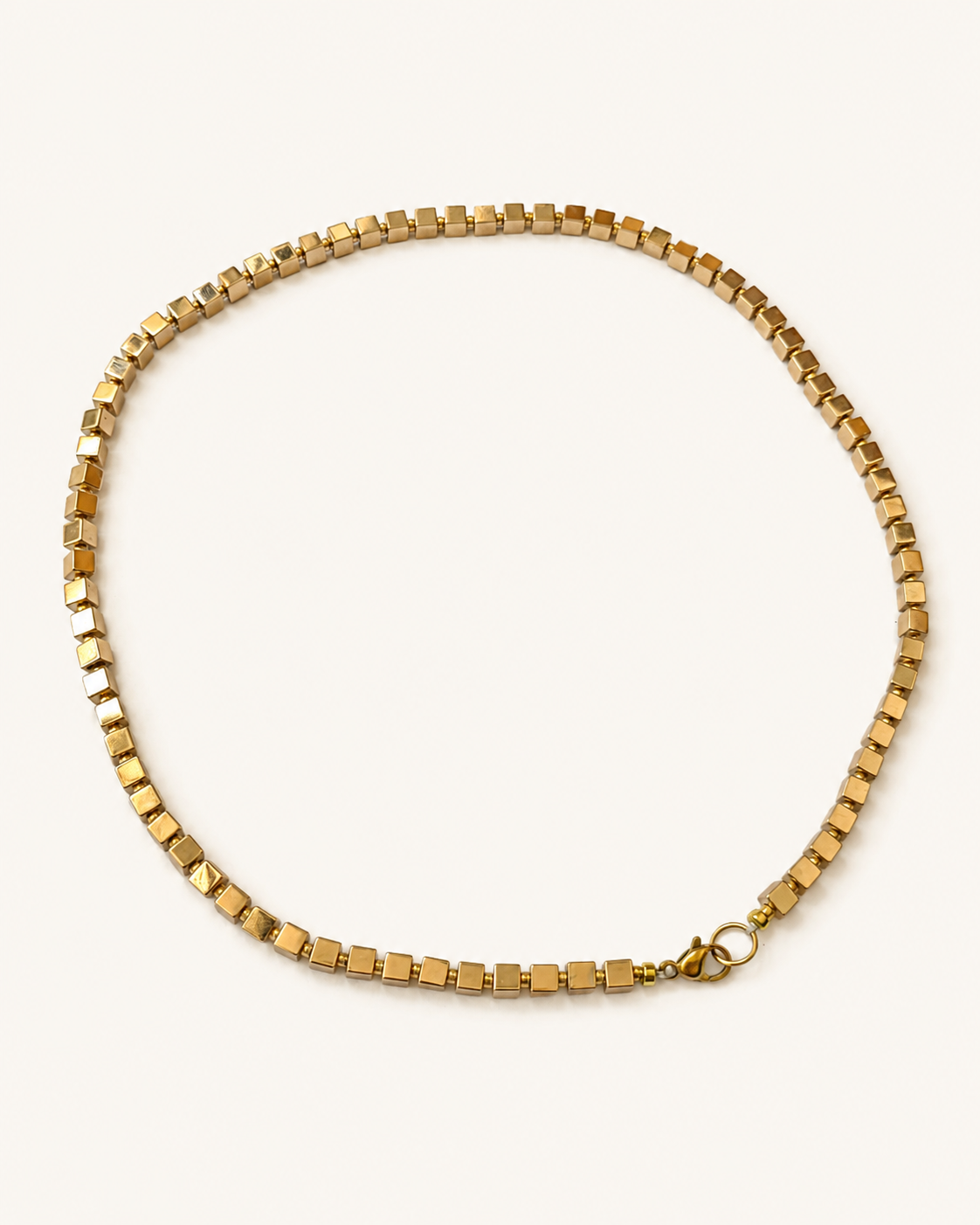 Angled full view of a gold-coloured hematite cube necklace on a warm ivory background