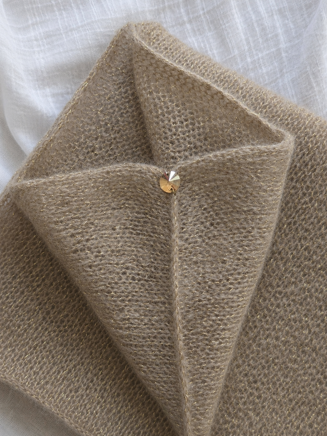 A close-up of a folded beige knit fabric item, likely a scarf or shawl, with a small gold heart-shaped pin or charm at the center, resting on a white textured fabric background.