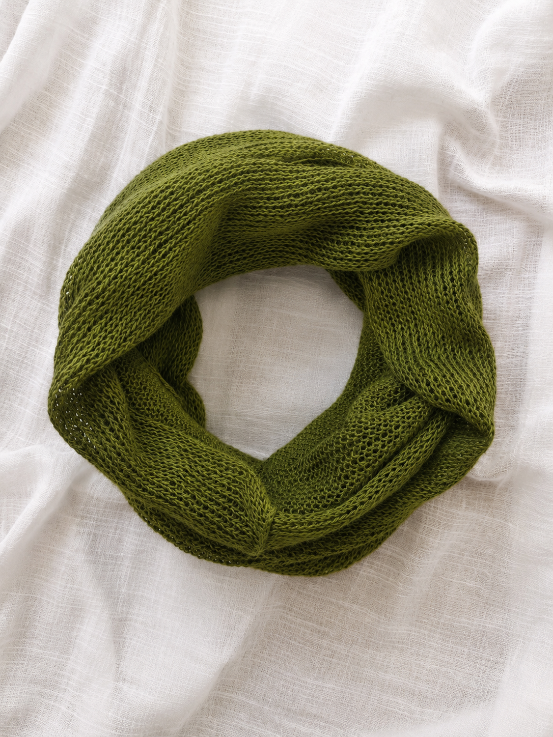 An olive green chunky-knit infinity scarf, styled in a loop on a textured white linen fabric background.