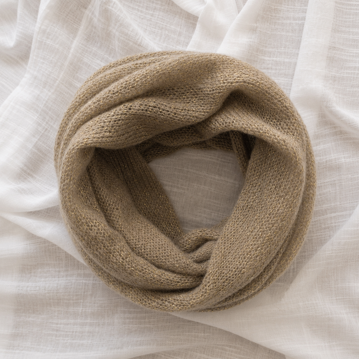A chunky knit infinity scarf in a warm taupe-beige color, styled in a loose coil on a draped white linen fabric background.