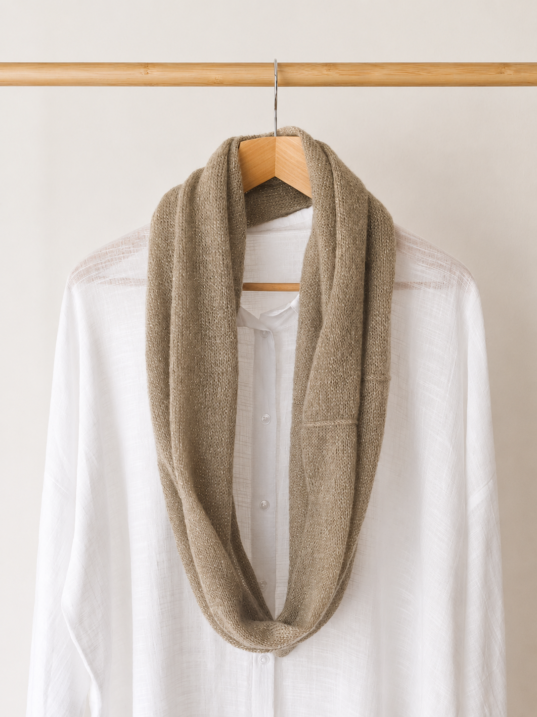 A thick, soft knit infinity scarf in a light taupe-beige color, draped over a white button-up shirt on a wooden hanger hanging from a wooden clothing rack.