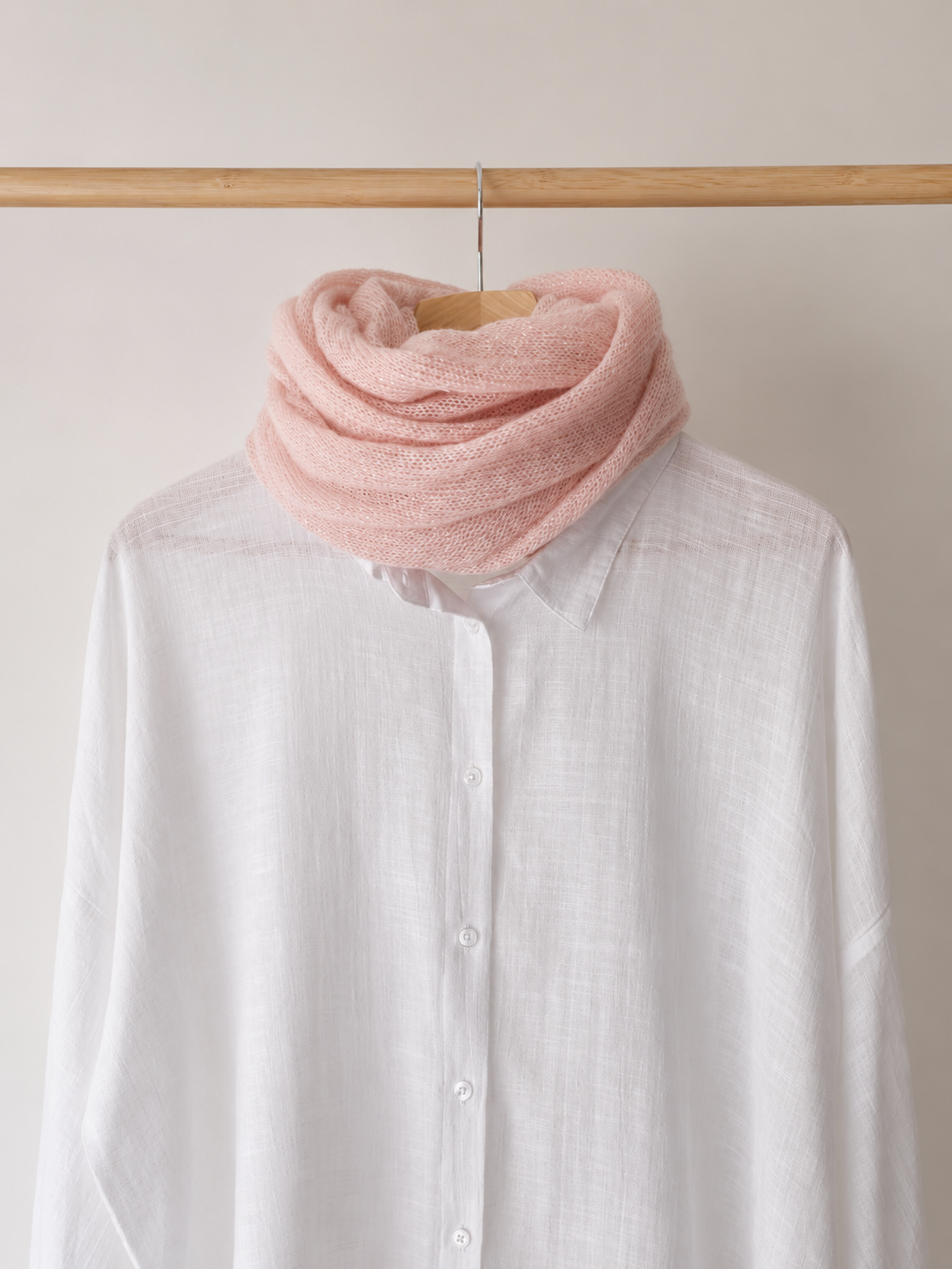 A soft dusty pink infinity scarf wrapped around a white linen button-up shirt, displayed on a wooden hanger hanging from a wooden clothing rack against a light beige wall.