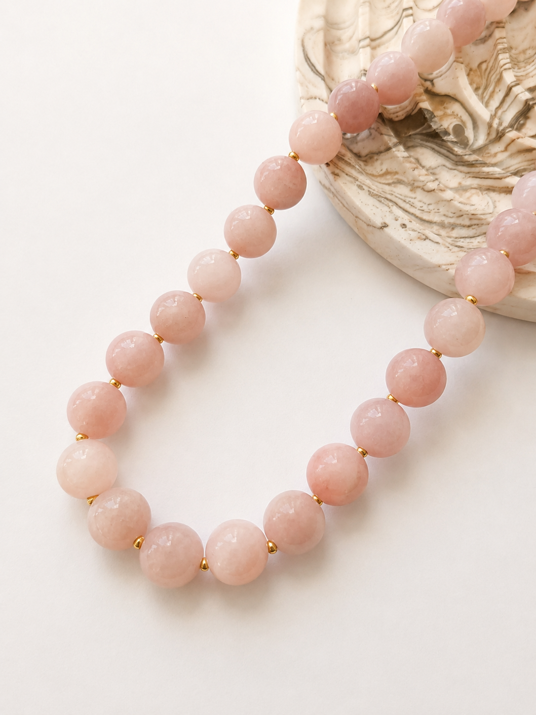Blush pink jadeite necklace styled beside a marbled decorative object on a warm ivory background.