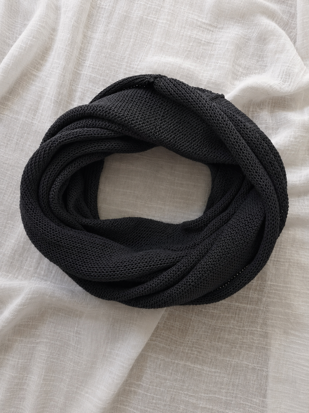 A black knit infinity scarf, coiled and laid flat on a textured off-white linen fabric background.