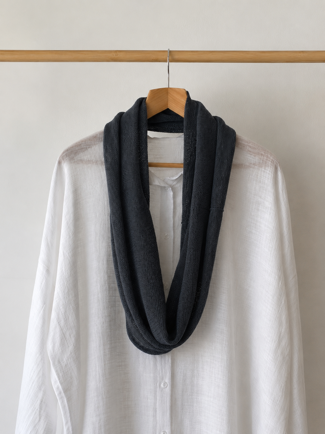 A dark charcoal gray infinity scarf draped over a white linen button-up shirt on a wooden hanger, hanging from a light wood clothing rack against a plain off-white wall.