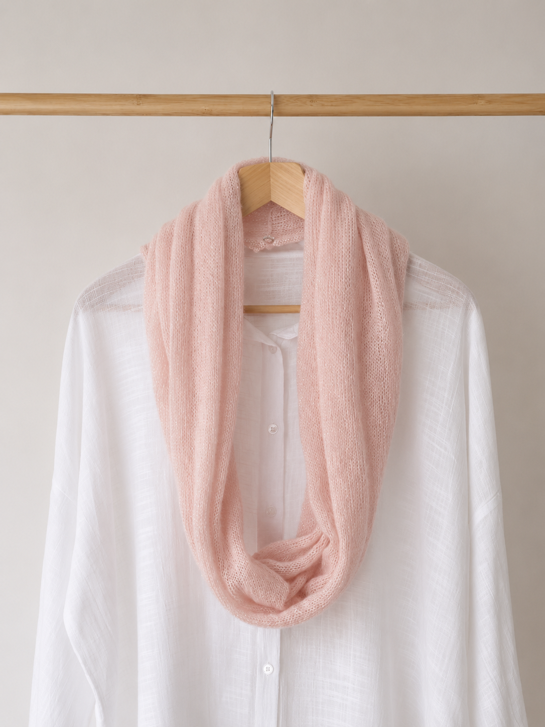 A soft blush pink infinity scarf draped over a white button-up shirt on a wooden hanger, hanging from a wooden clothes rack against a light beige wall.