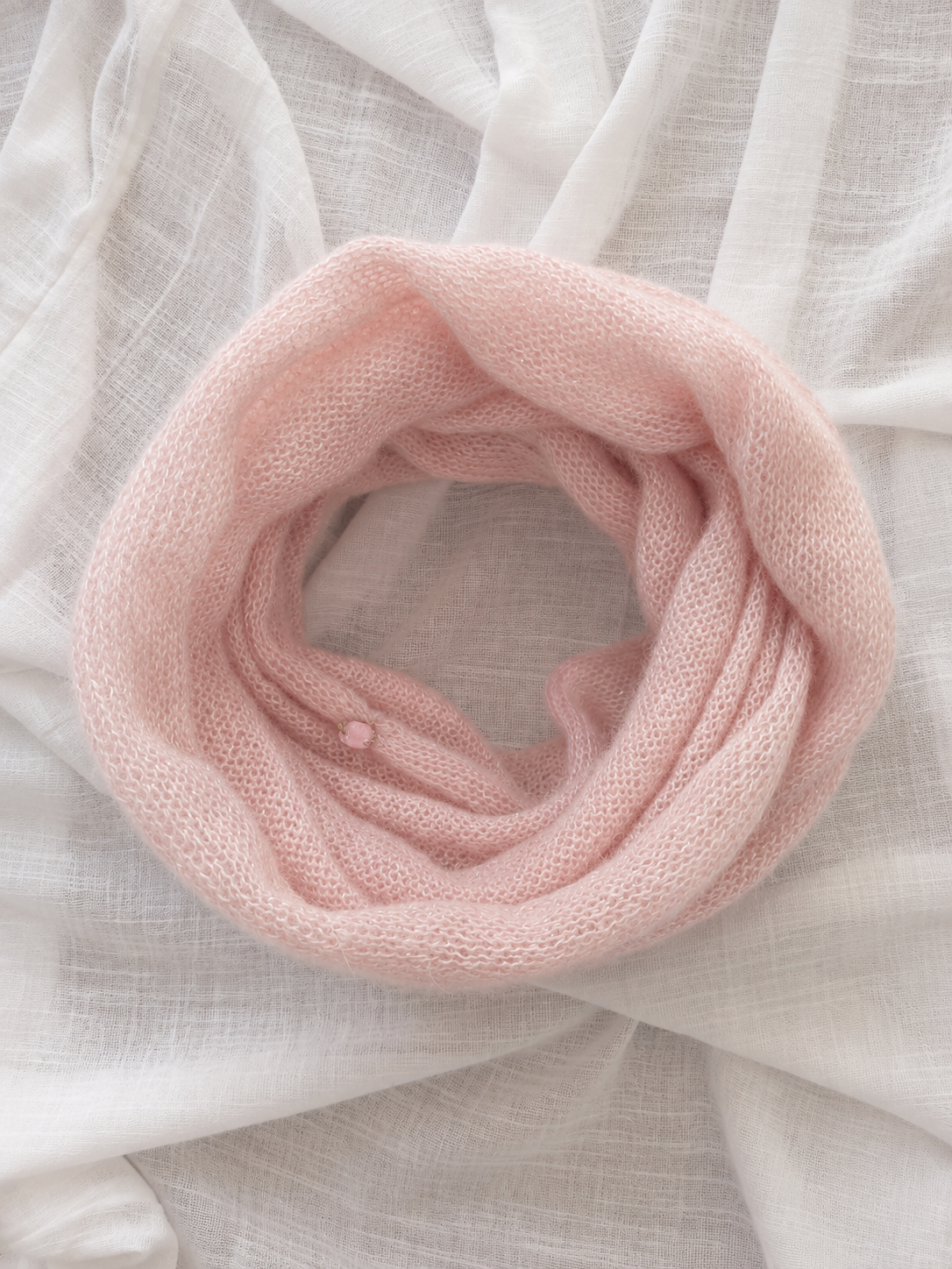 A soft, light pink knit infinity scarf arranged in a loose loop on a draped white fabric background.