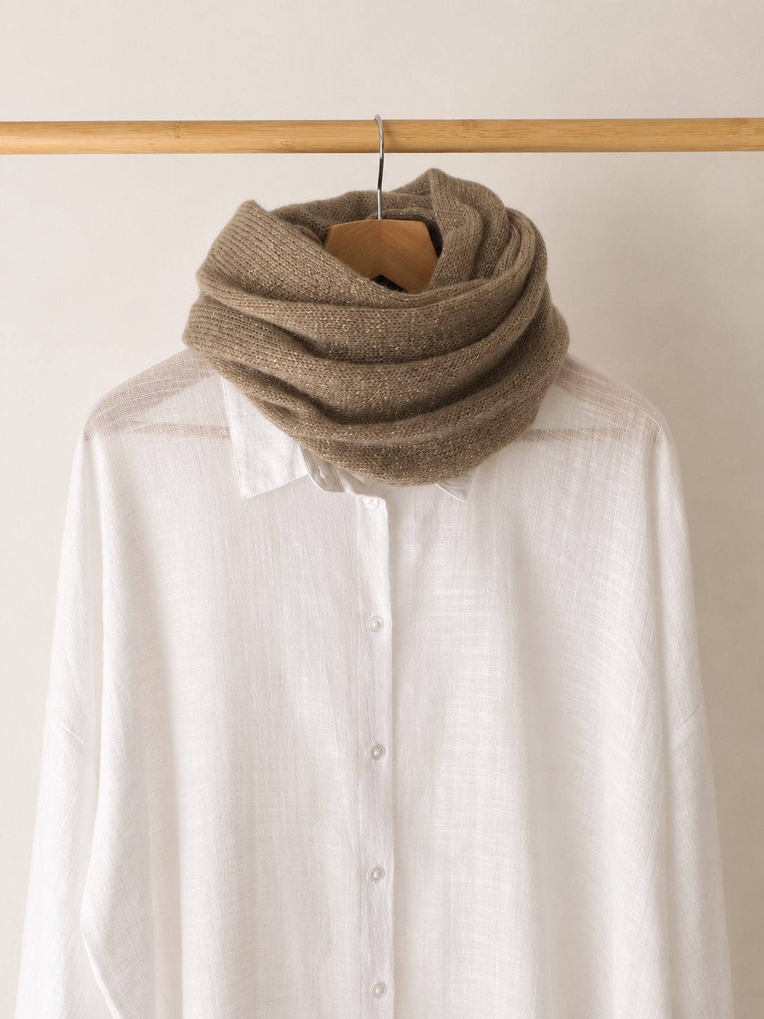 A thick, knitted brown infinity scarf wrapped around a white linen button-up shirt, hanging on a wooden hanger from a light wood clothing rack against a plain off-white wall.