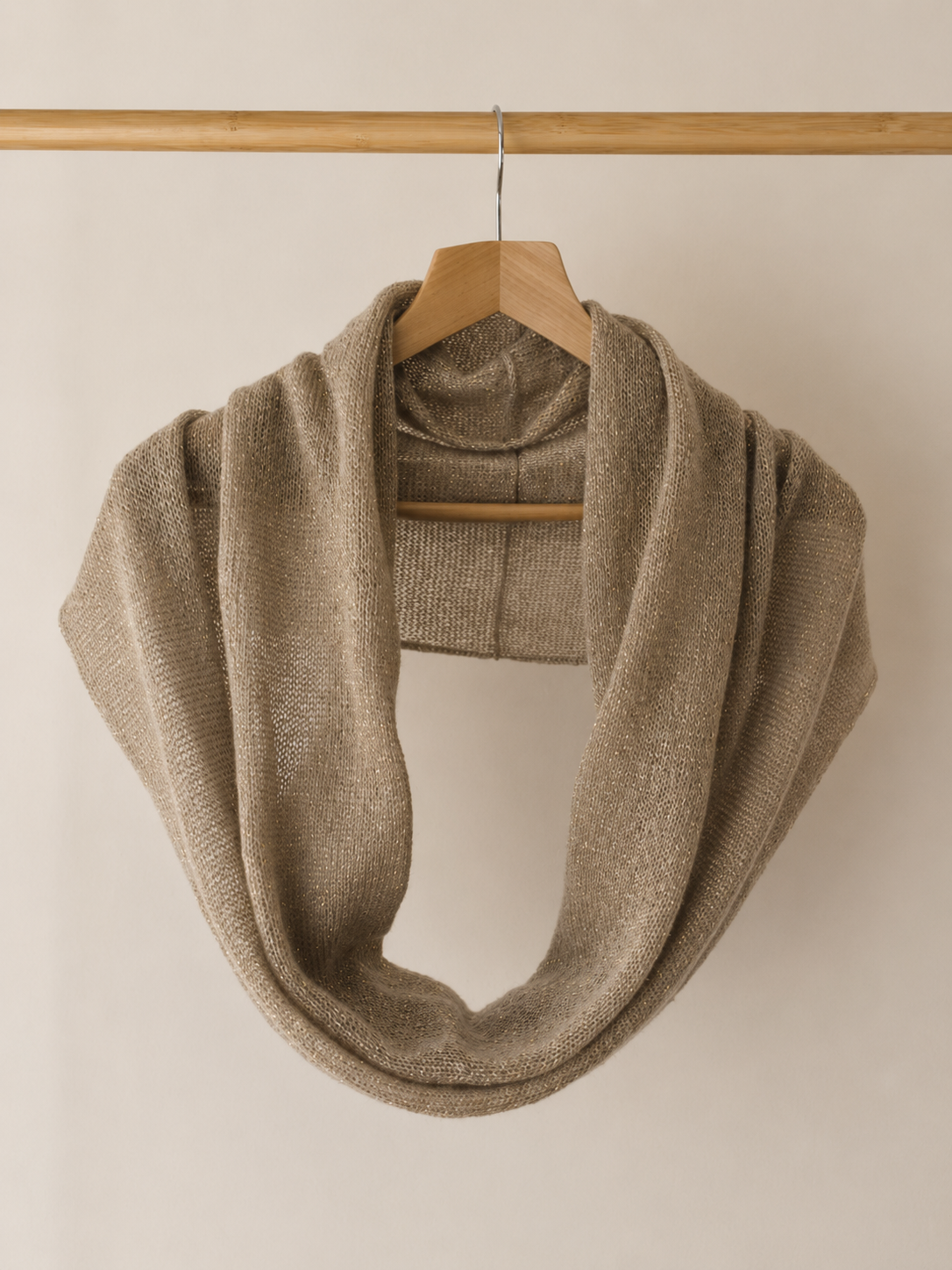 A chunky knit infinity scarf in a warm taupe-beige color, draped over a wooden hanger hanging from a wooden rod against a plain light beige wall.